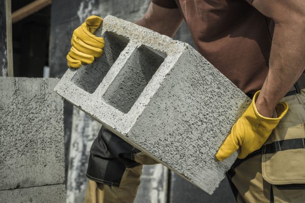 A construction worker carrying a stack of masonry blocks and walking toward a brick wall